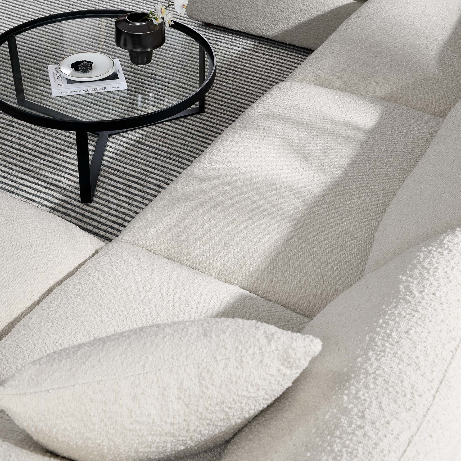 Ivory boucle sectional sofa with matching pillow, round glass-top black metal coffee table on striped grey rug.