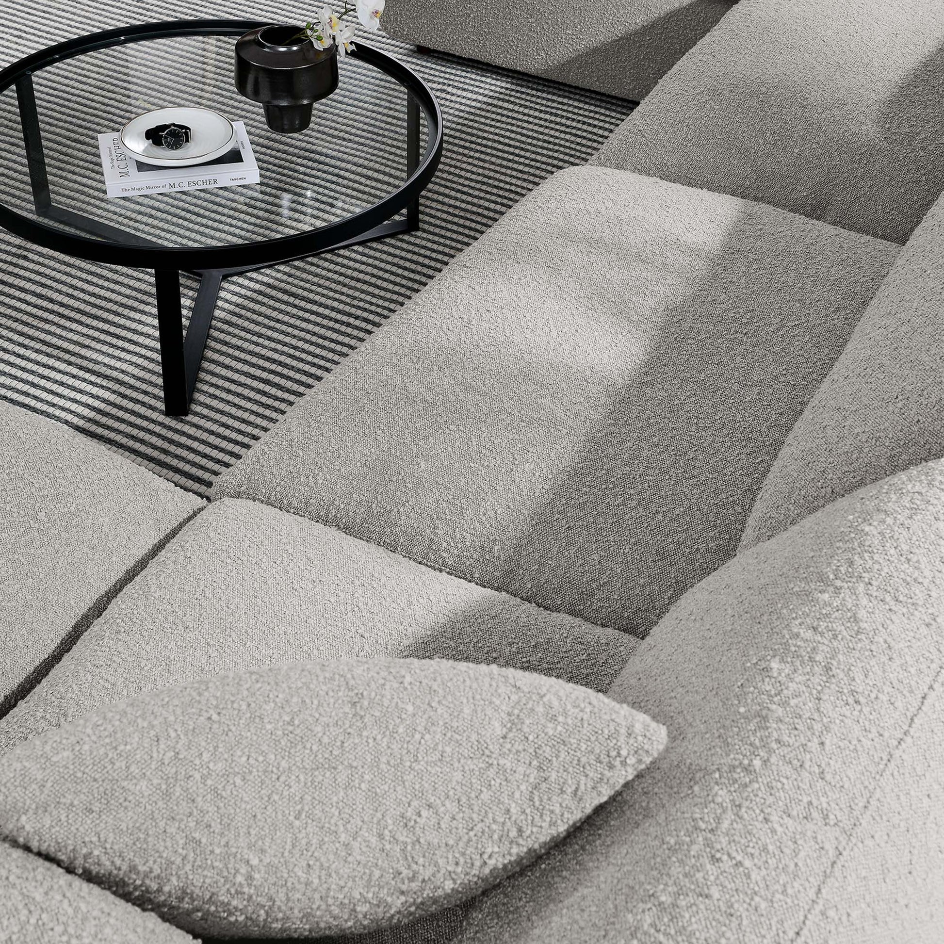 Light gray boucle sectional sofa with cushions, round glass coffee table on black metal legs, striped rug, magazine on table.