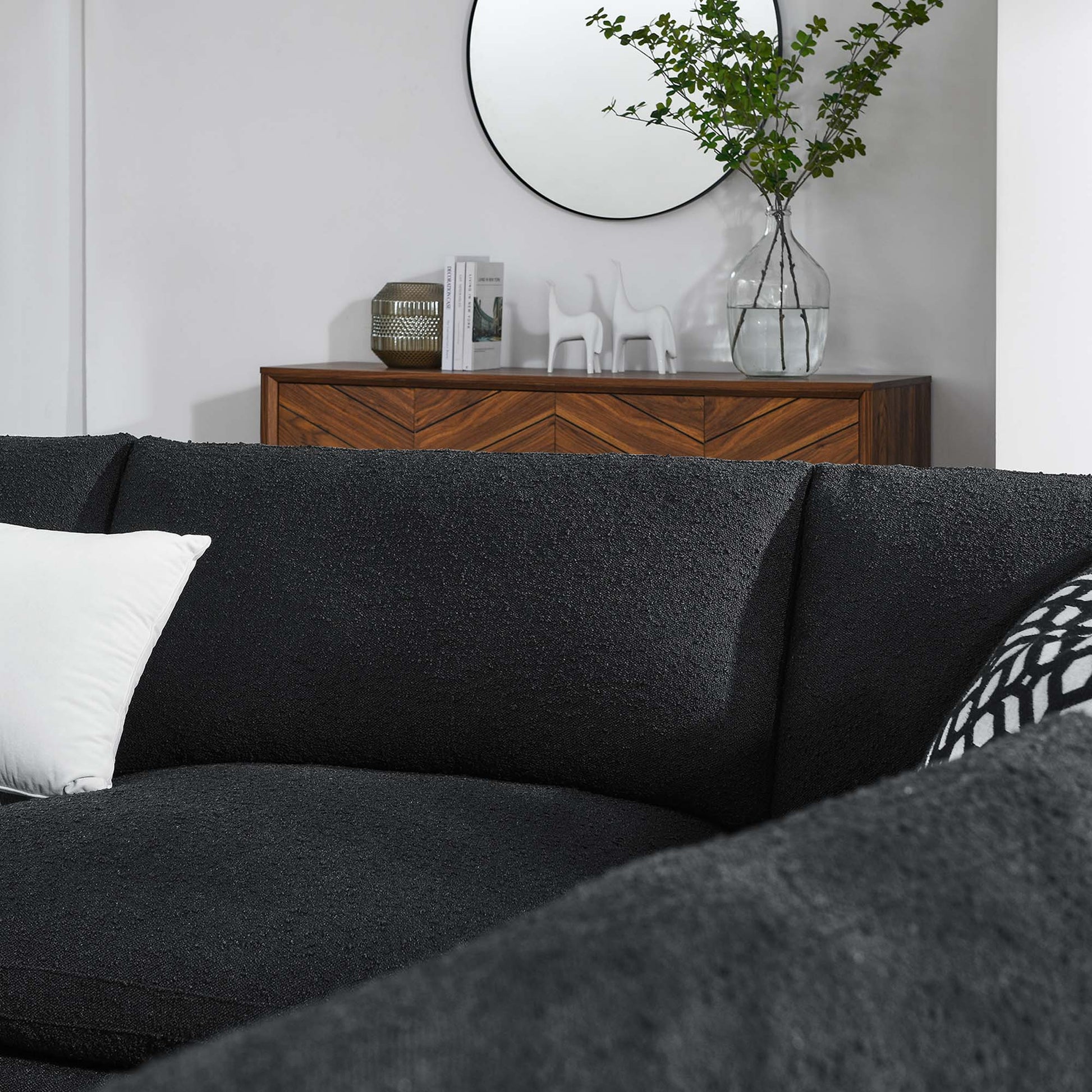 Charcoal fabric sectional sofa with white pillow, wooden sideboard, round mirror, vase of greenery, and white decorative figurines.