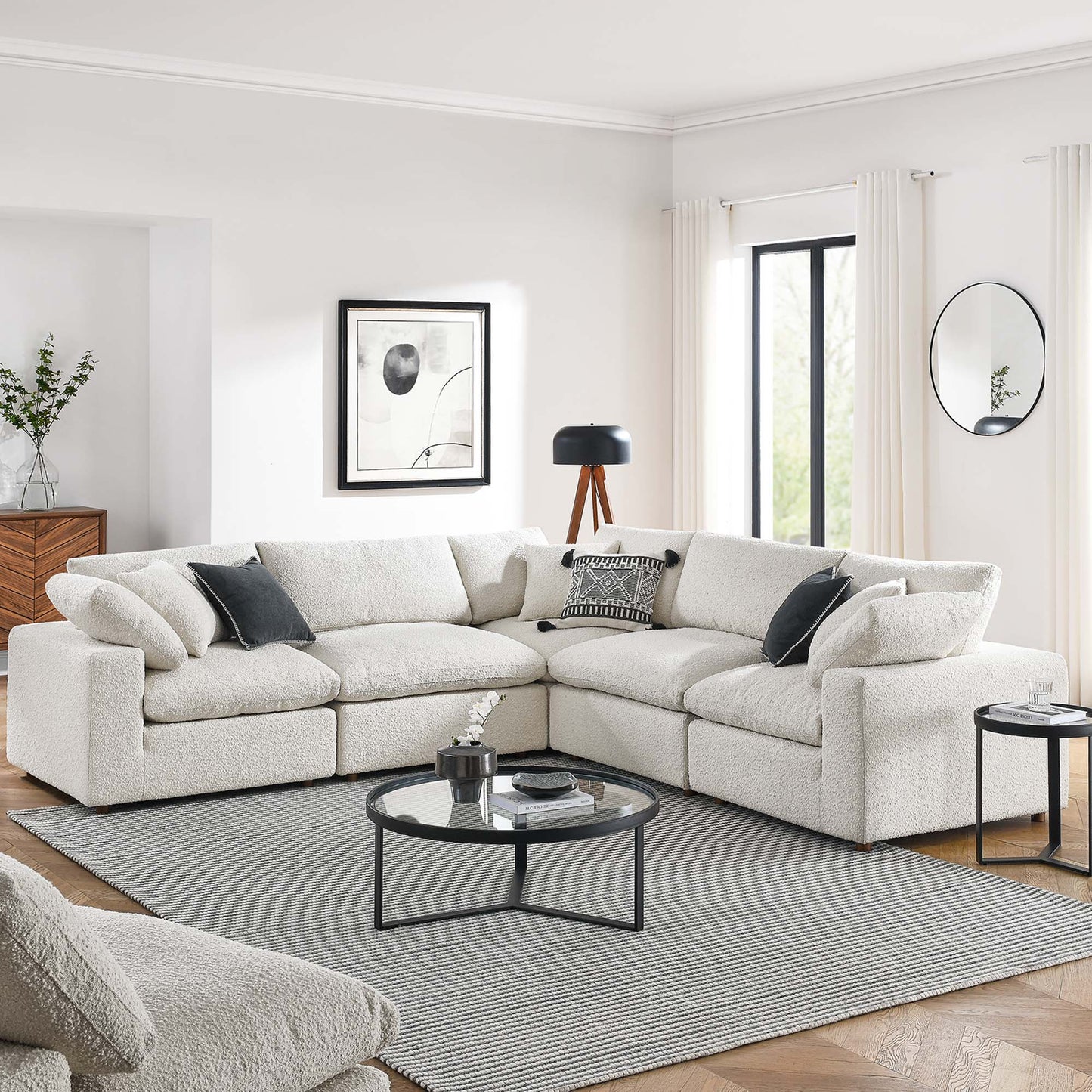 Ivory L-shaped boucle sectional sofa with dark throw pillows, round glass coffee table on gray rug in modern living room.