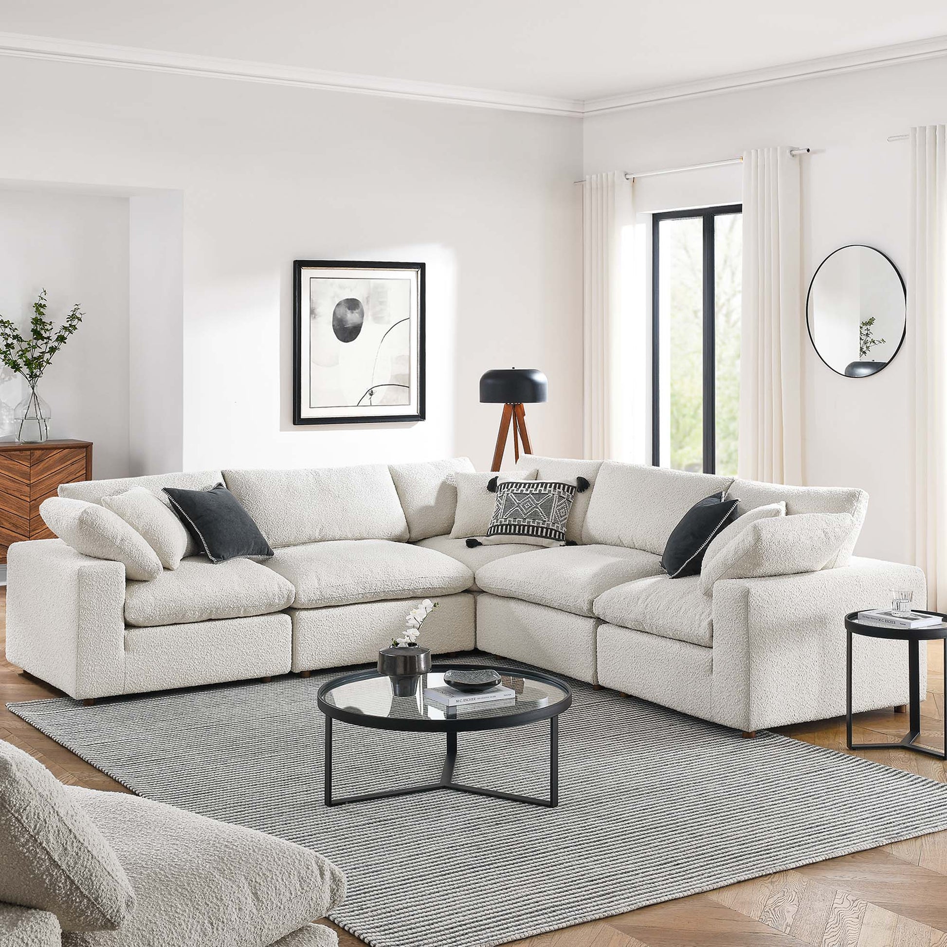 Ivory L-shaped boucle sectional sofa with dark throw pillows, round glass coffee table on gray rug in modern living room.