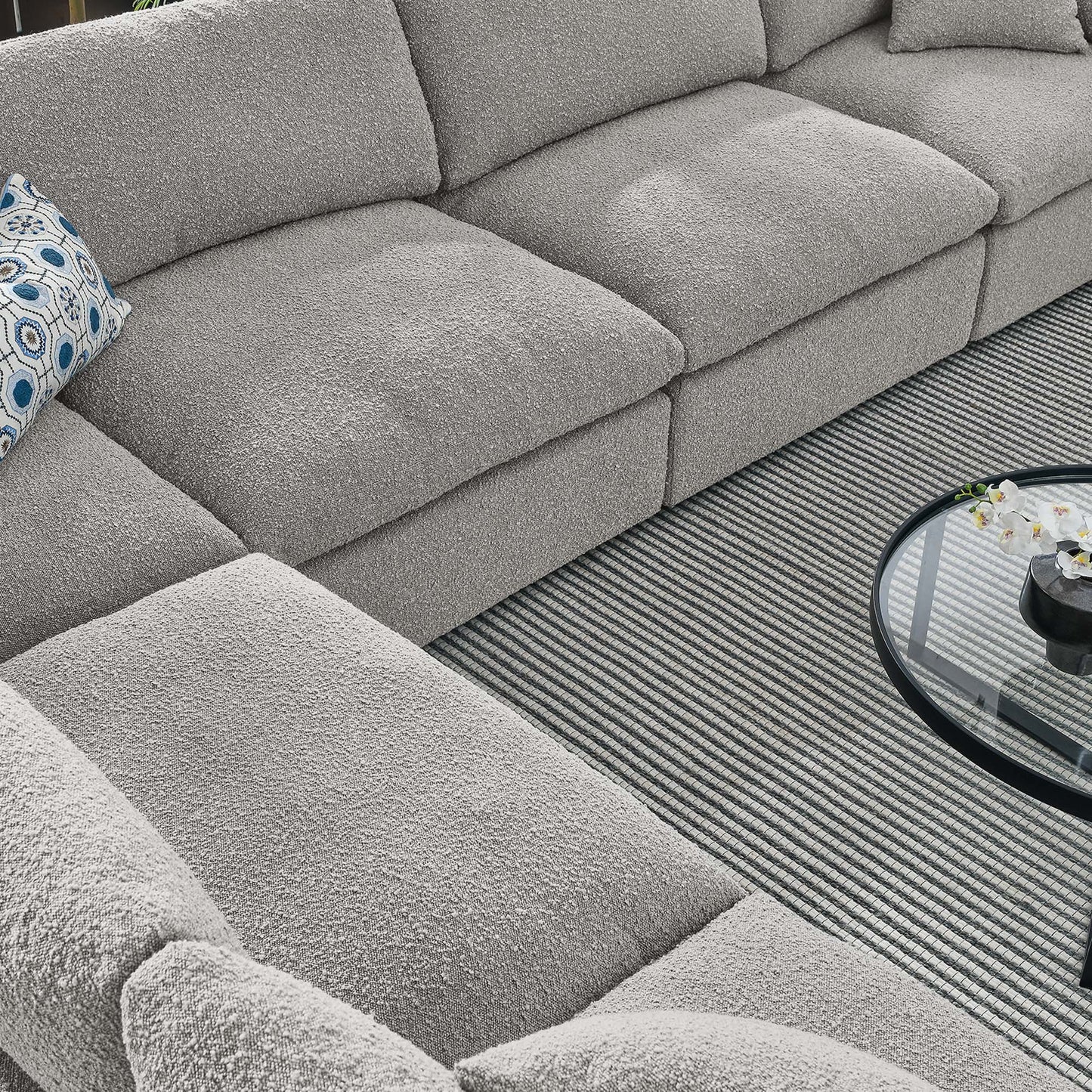 L-shaped gray boucle sectional sofa with a blue patterned pillow and round glass coffee table on a striped rug.
