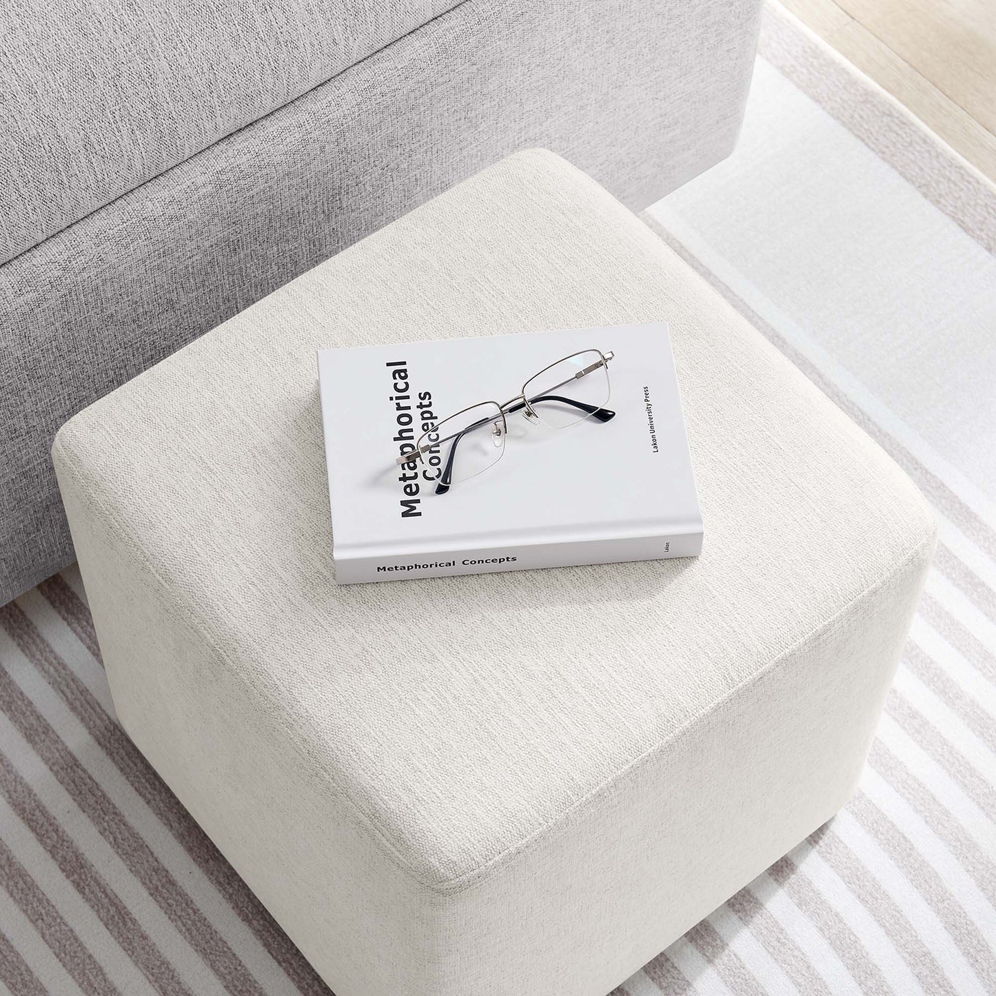 Cream fabric ottoman with a Metaphorical Concepts book and metal glasses on top in a neutral living room.