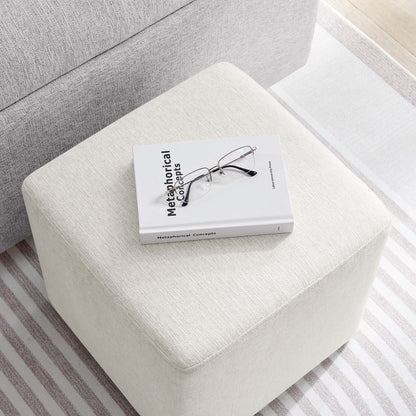 Cream fabric ottoman with a Metaphorical Concepts book and metal glasses on top in a neutral living room.