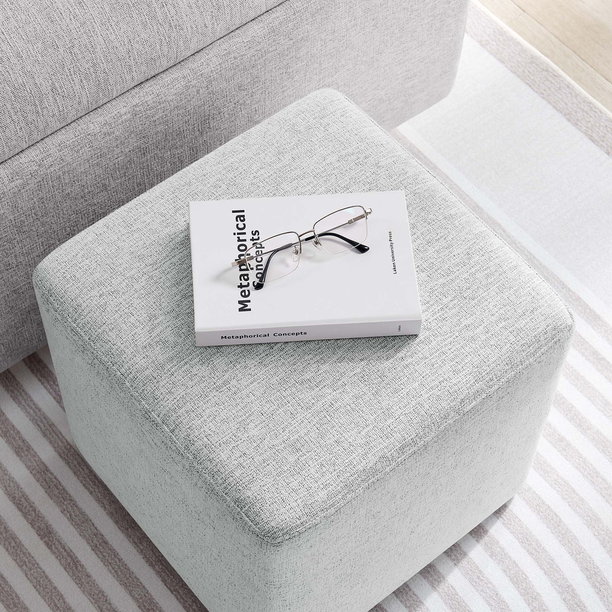 Light gray fabric ottoman with white Metaphorical Concepts book and metal glasses on top; minimalist living room.
