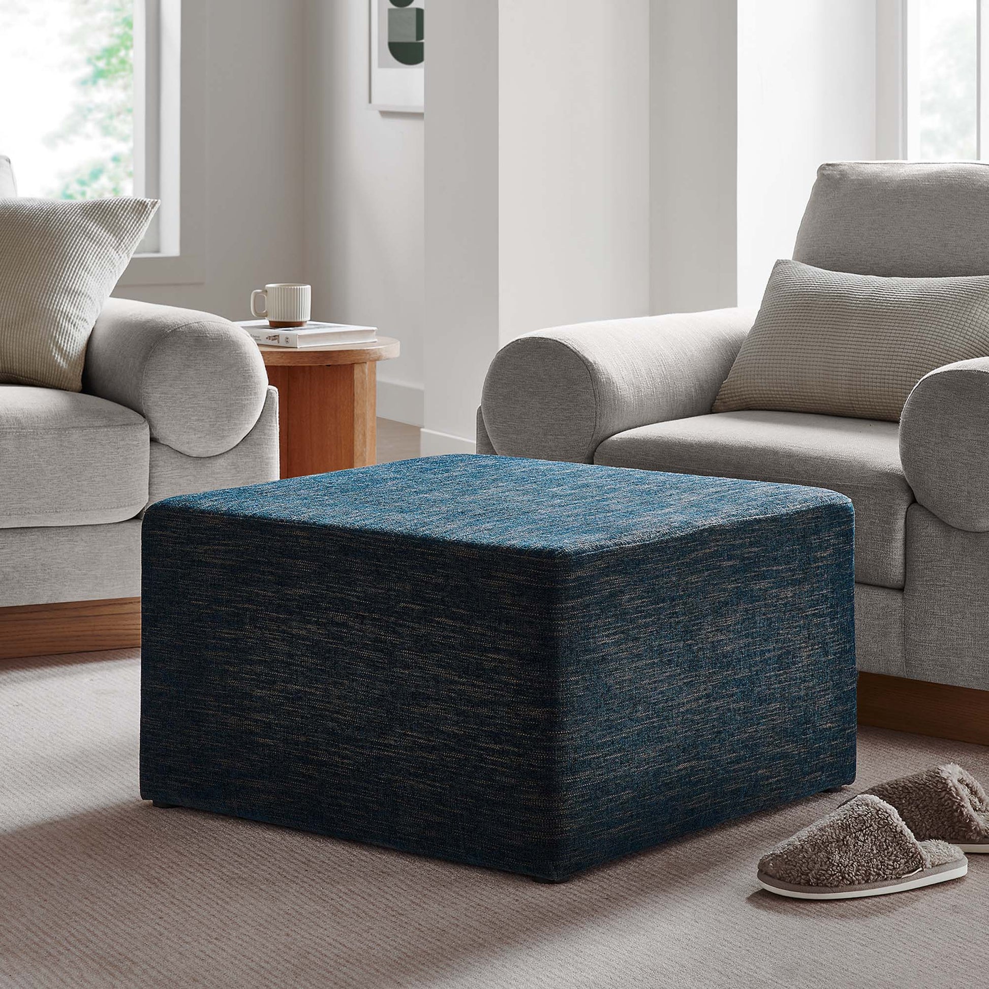 Blue textured fabric 28-inch square ottoman between two gray sofas in a contemporary living room with slippers nearby.