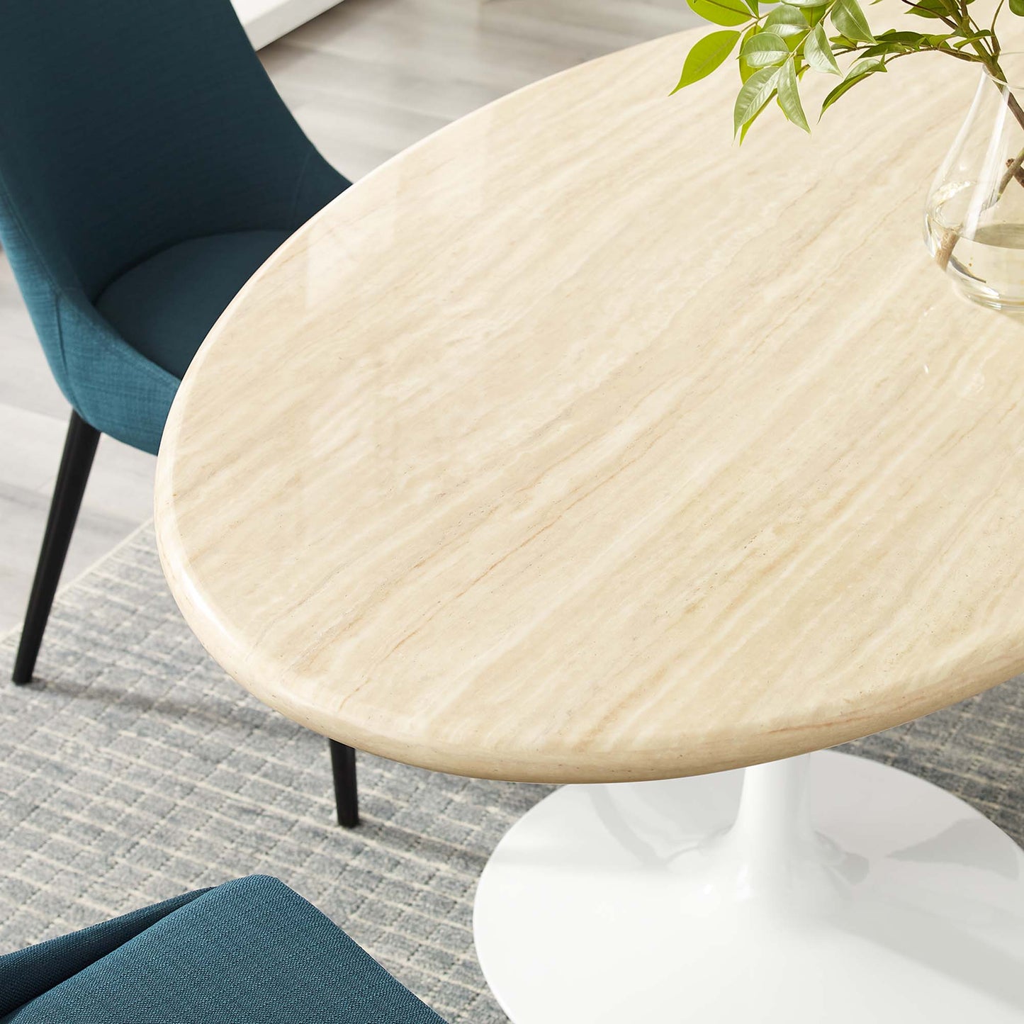 Oval faux travertine dining table with teal upholstered dining chairs and a glass vase with greenery.