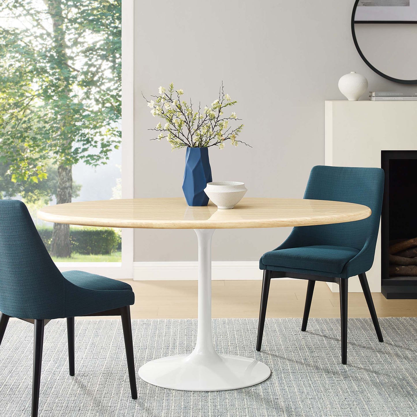 Oval faux travertine dining table with white pedestal base, teal chairs & blue geometric vase with branches in a bright living room.