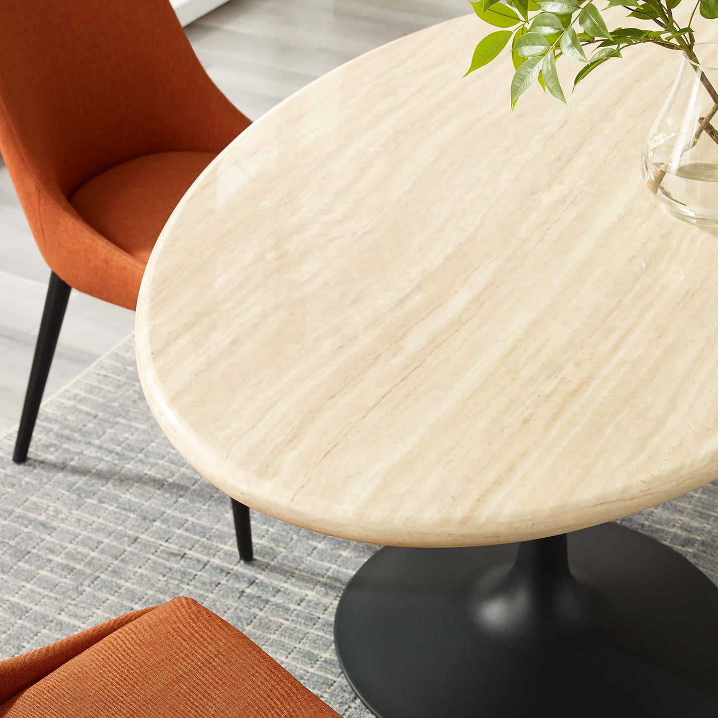 Oval faux travertine dining table with a pedestal base, orange upholstered chairs, and a glass vase with greenery.