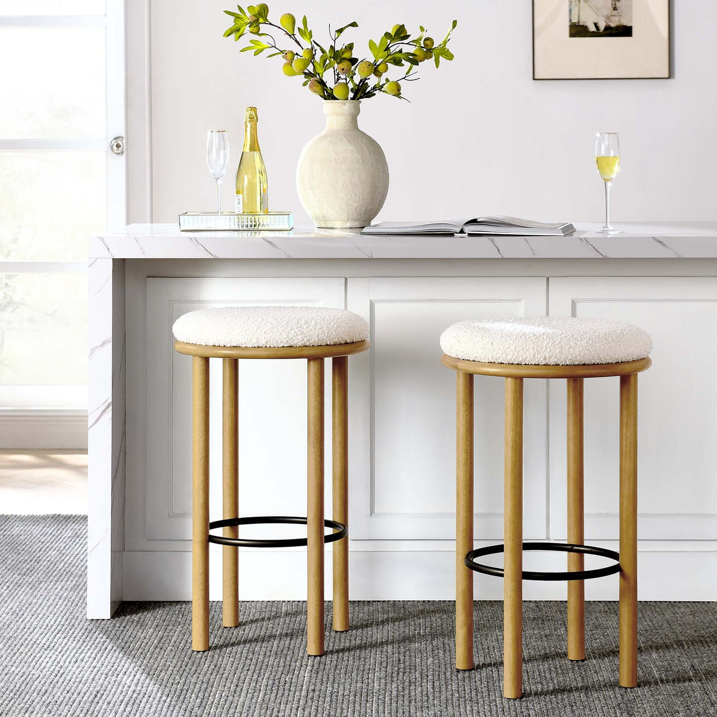 Two ivory-cushioned oak bar stools with black footrests at a white kitchen island, with vase and champagne.