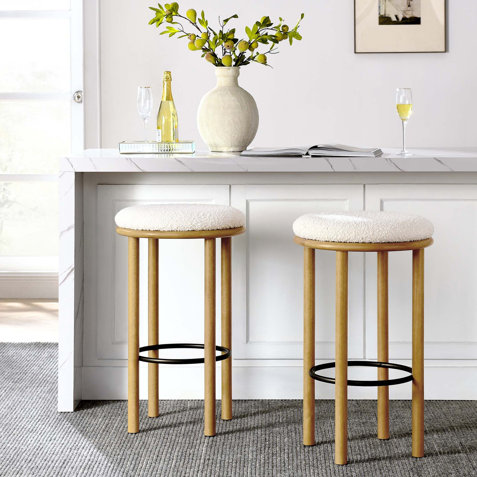 Two ivory-cushioned oak bar stools with black footrests at a white kitchen island, with vase and champagne.