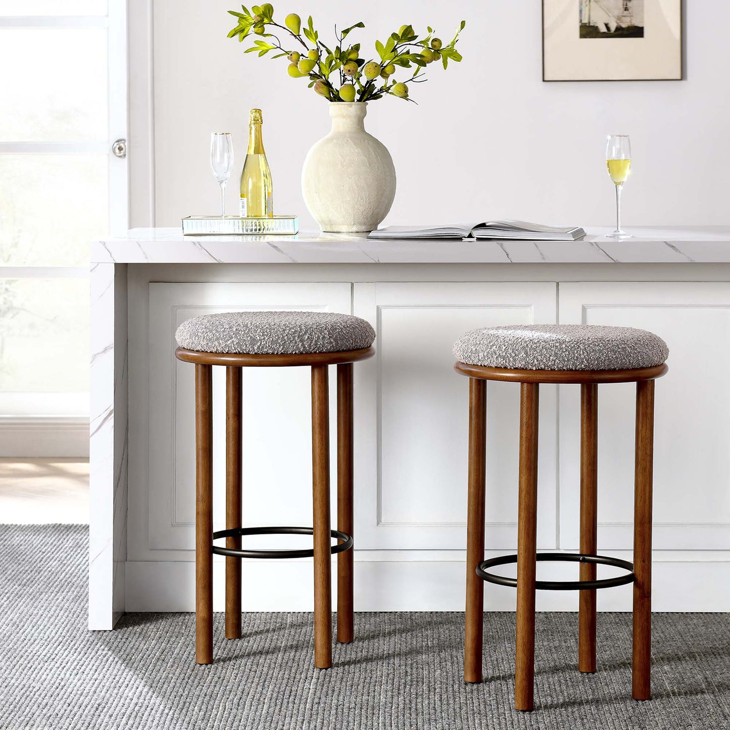 Two wooden bar stools with taupe fabric seats at a white kitchen island, with a vase of greenery, champagne bottle, and glasses.
