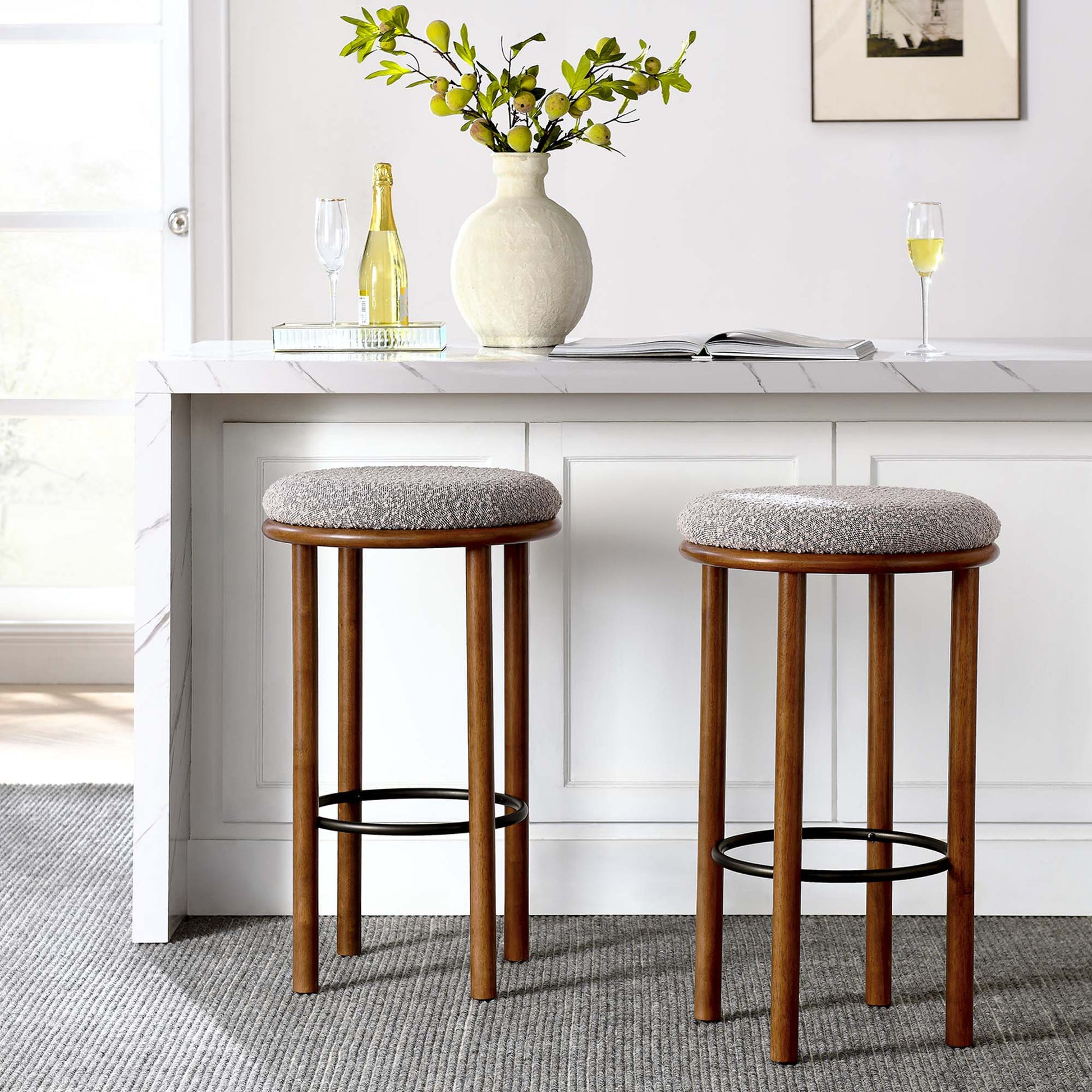 Two wooden bar stools with taupe fabric seats at a white kitchen island, with a vase of greenery, champagne bottle, and glasses.