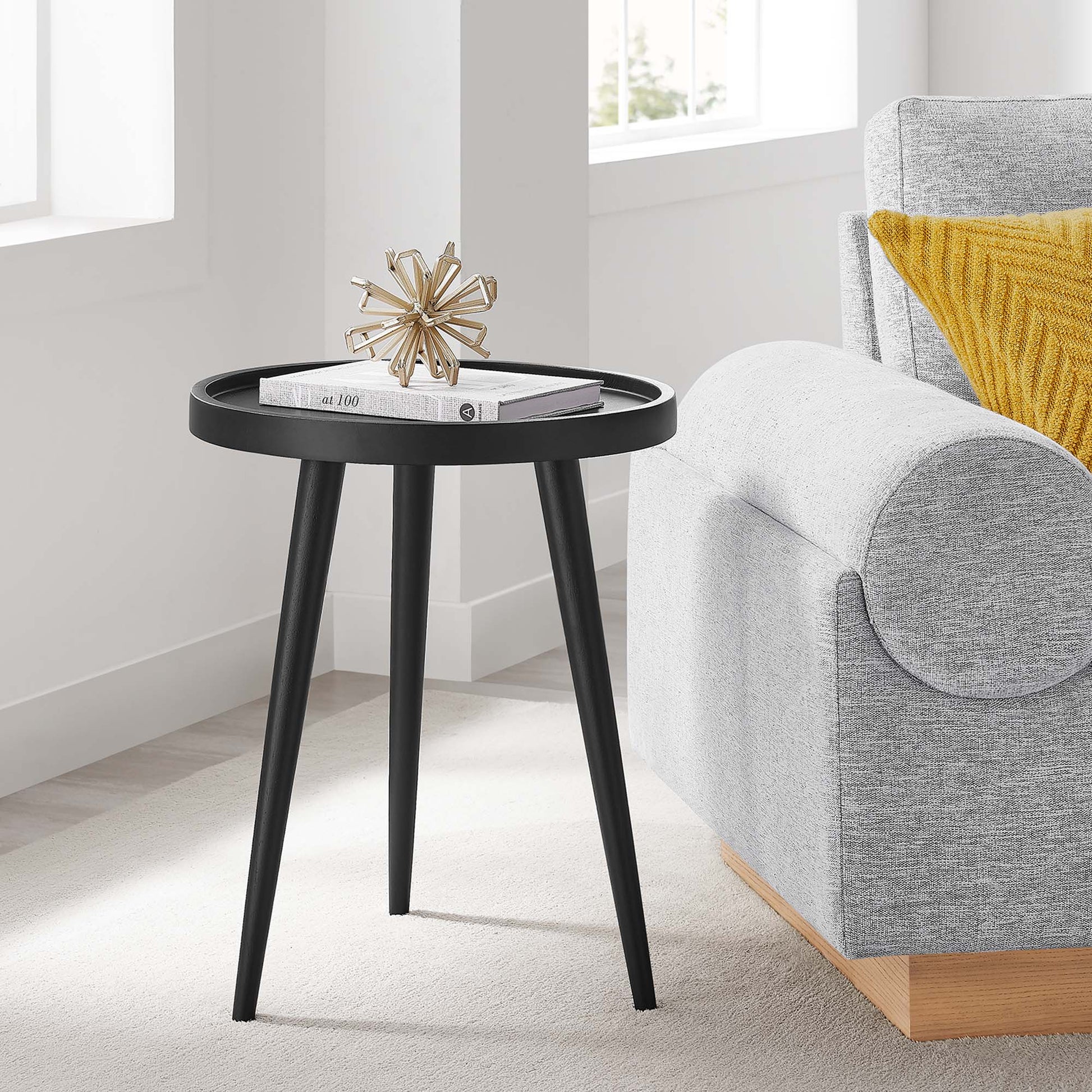 Black round tripod side table with three tapered legs; magazines and a gold starburst sculpture on top beside a gray sofa.