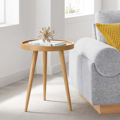 Natural wood round side table with three tapered legs, magazines on top, in a bright living room beside a gray sofa.