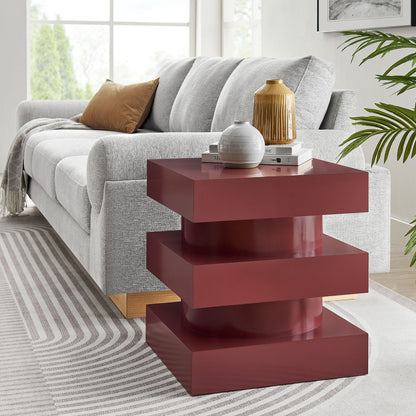 Glossy burgundy stacked end table with three wide platforms beside a light gray sofa; decor books and ceramic vases on top.