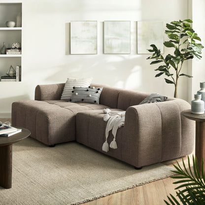 Brown left-facing upholstered fabric modular sofa with chaise, cushions, and tassel throw in a bright living room with abstract art, a plant, and a round coffee table.