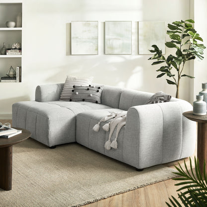 Light gray fabric sectional with chaise in a modern living room; tassel throw, striped pillow, beige rug, abstract wall art.