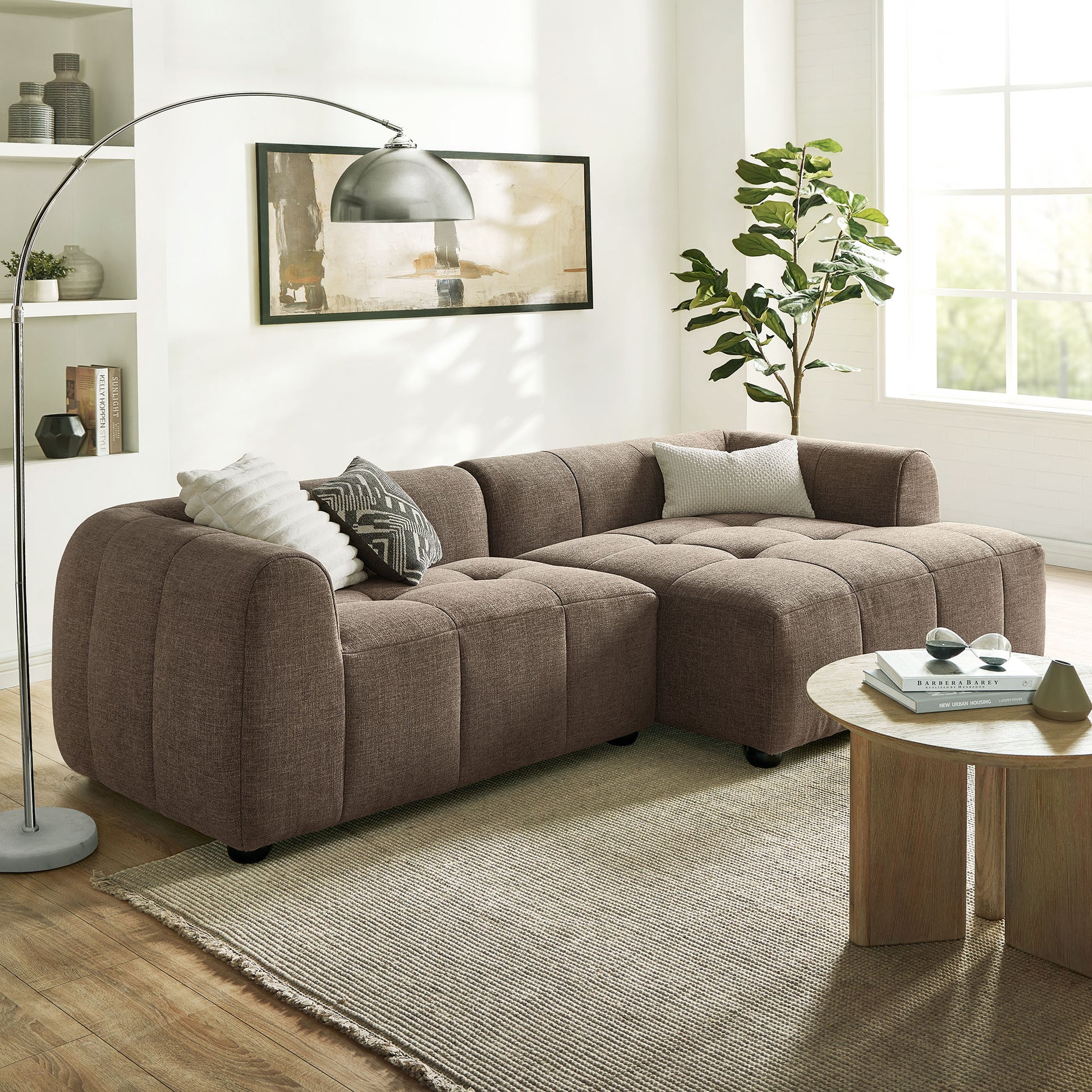 Brown modular fabric sectional sofa in an L-shape with tufted cushions, with round wood coffee table and an arc lamp.