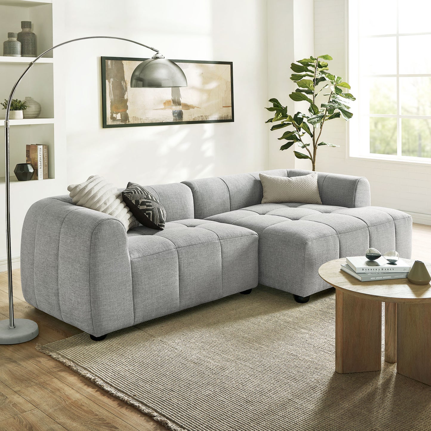 Light gray fabric modular sectional sofa in an L-shape with tufted cushions; contemporary living room with a round wooden coffee table & plant.