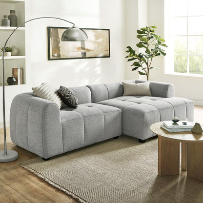 Light gray fabric modular sectional sofa in an L-shape with tufted cushions; contemporary living room with a round wooden coffee table & plant.