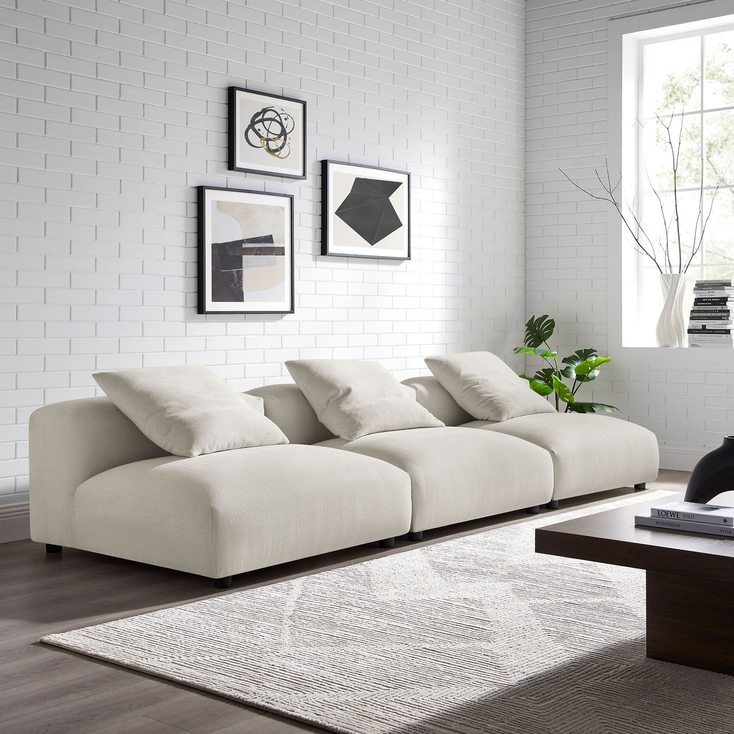 Gray fabric modular sofa with three cushions in a modern living room, white brick wall & abstract framed art, light rug, coffee table.