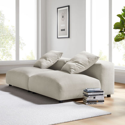 Brown modular upholstered fabric sofa with chaise and two large cushions in a bright living room; stacked books beside.
