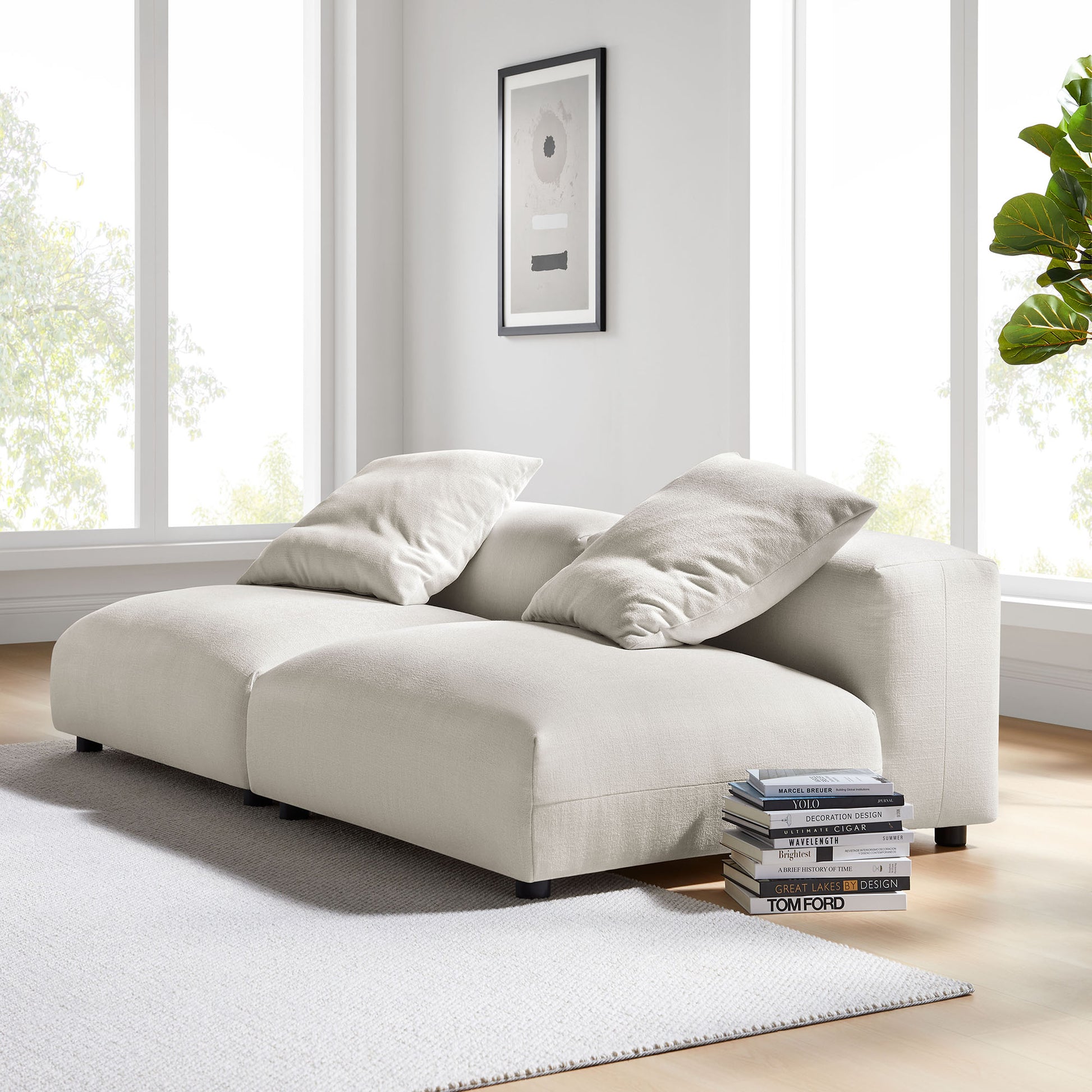 Gray fabric modular sofa with chaise, two large cushions, in a bright sunlit living room with a stack of books and framed art.