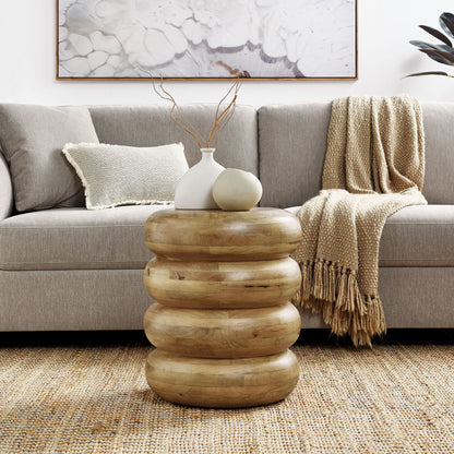 Natural wood stacked round end table with white vase, beige orb, twig stems, beside a beige sofa on a woven rug