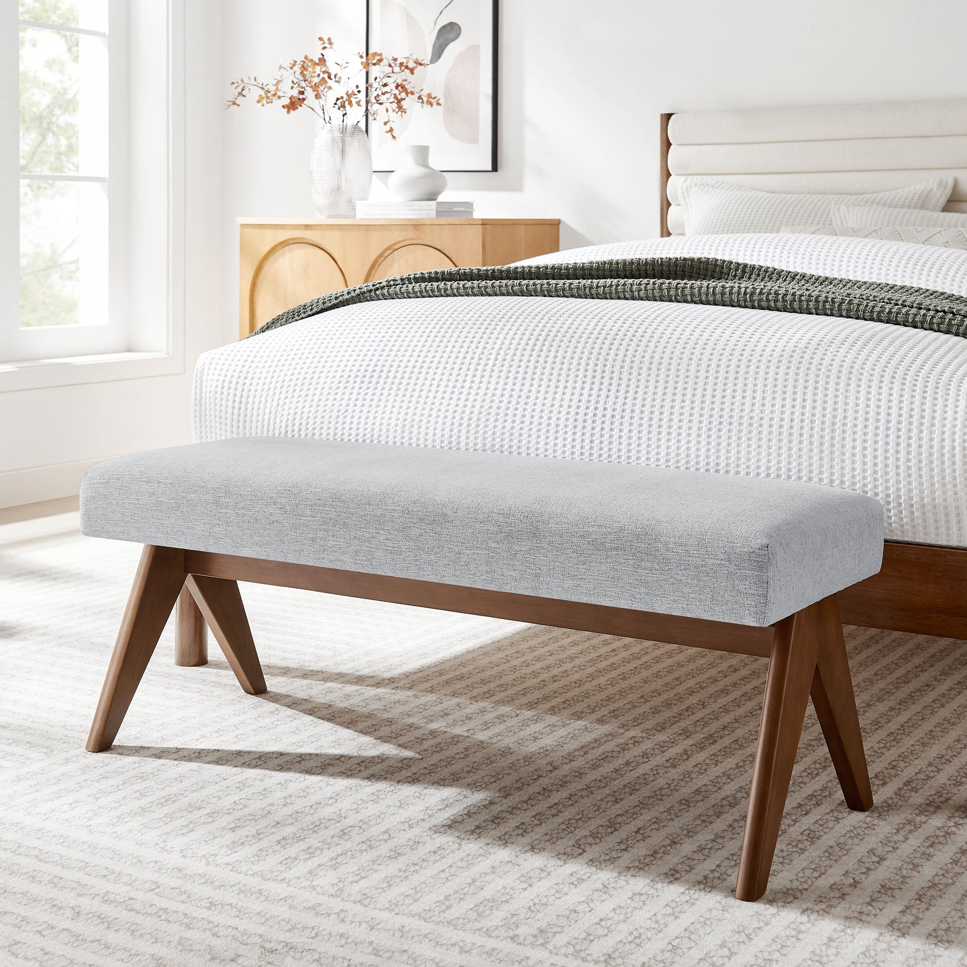 Light gray upholstered bench with angled wooden legs at foot of white bed in a bright, serene bedroom.