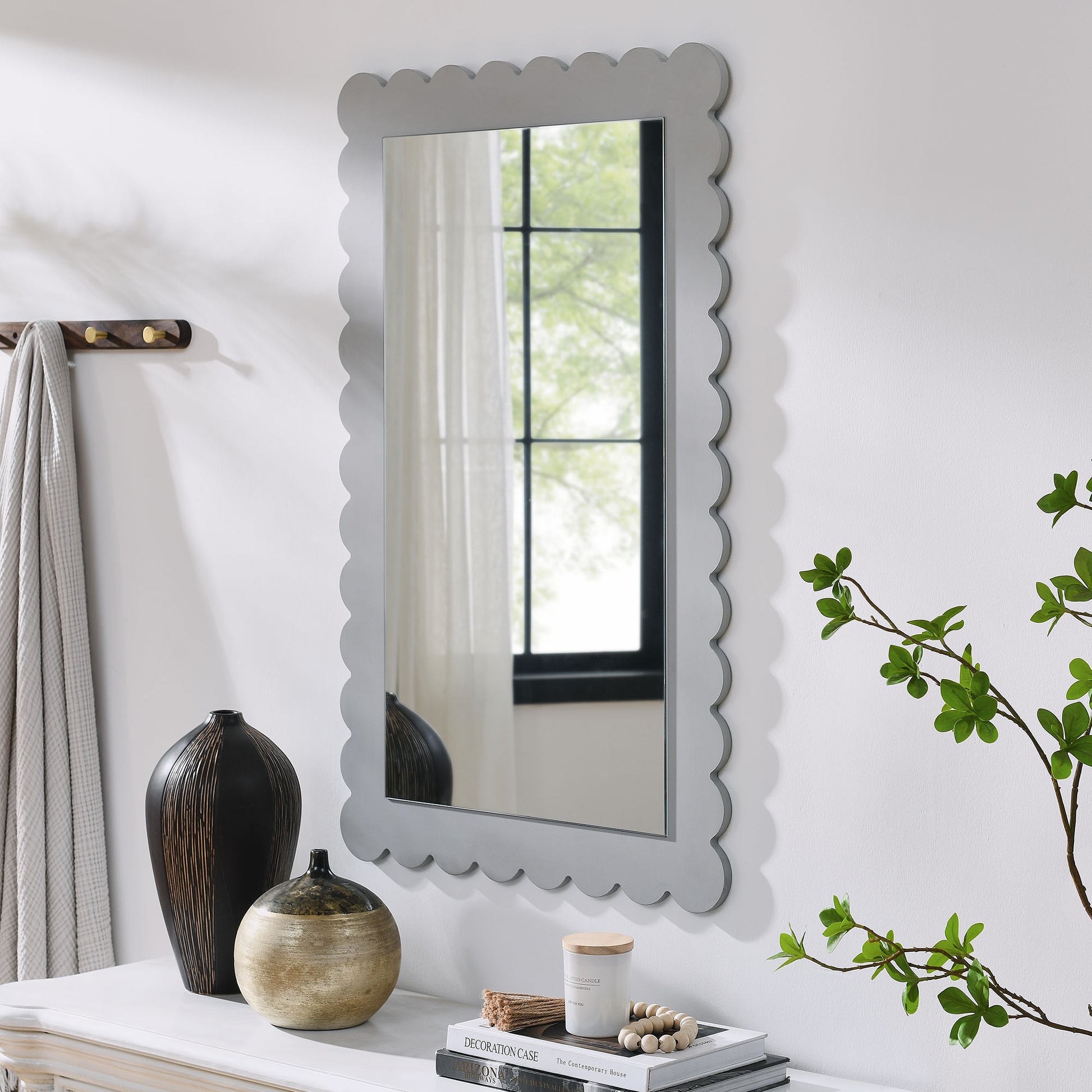 Scalloped gray-framed rectangle wall mirror over a white console with black vase, beige pot, candle, books, beads and greenery.