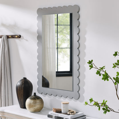 Scalloped gray-framed rectangle wall mirror over a white console with black vase, beige pot, candle, books, beads and greenery.