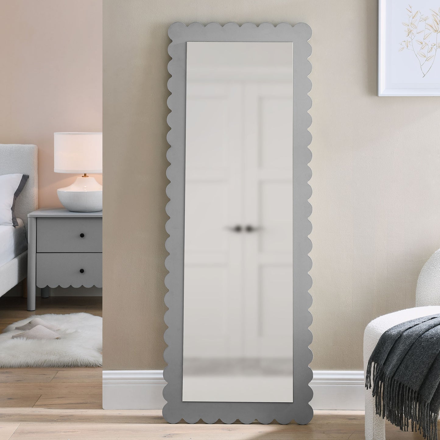 Tall full-length mirror with scalloped gray frame leaning against beige bedroom wall.