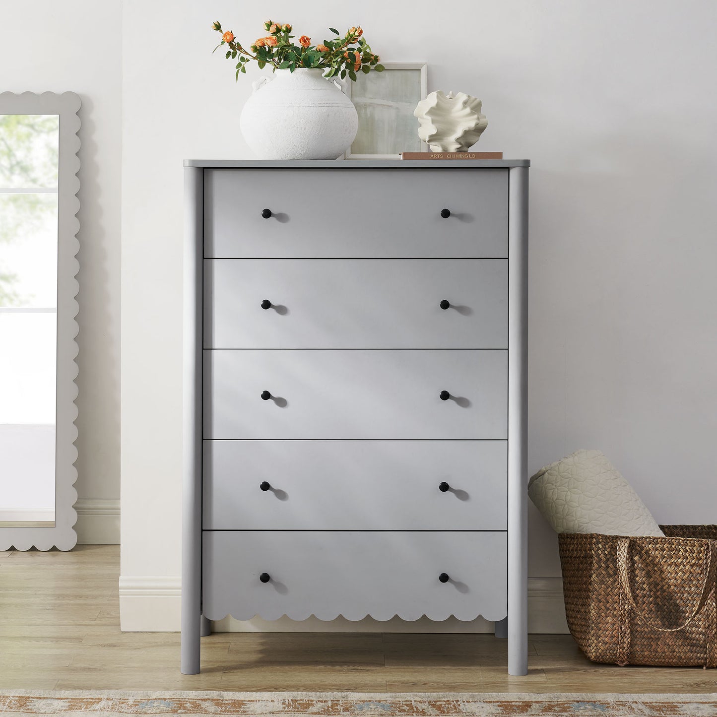 Gray five-drawer chest with scalloped apron and black knobs, topped by a white vase with orange flowers.