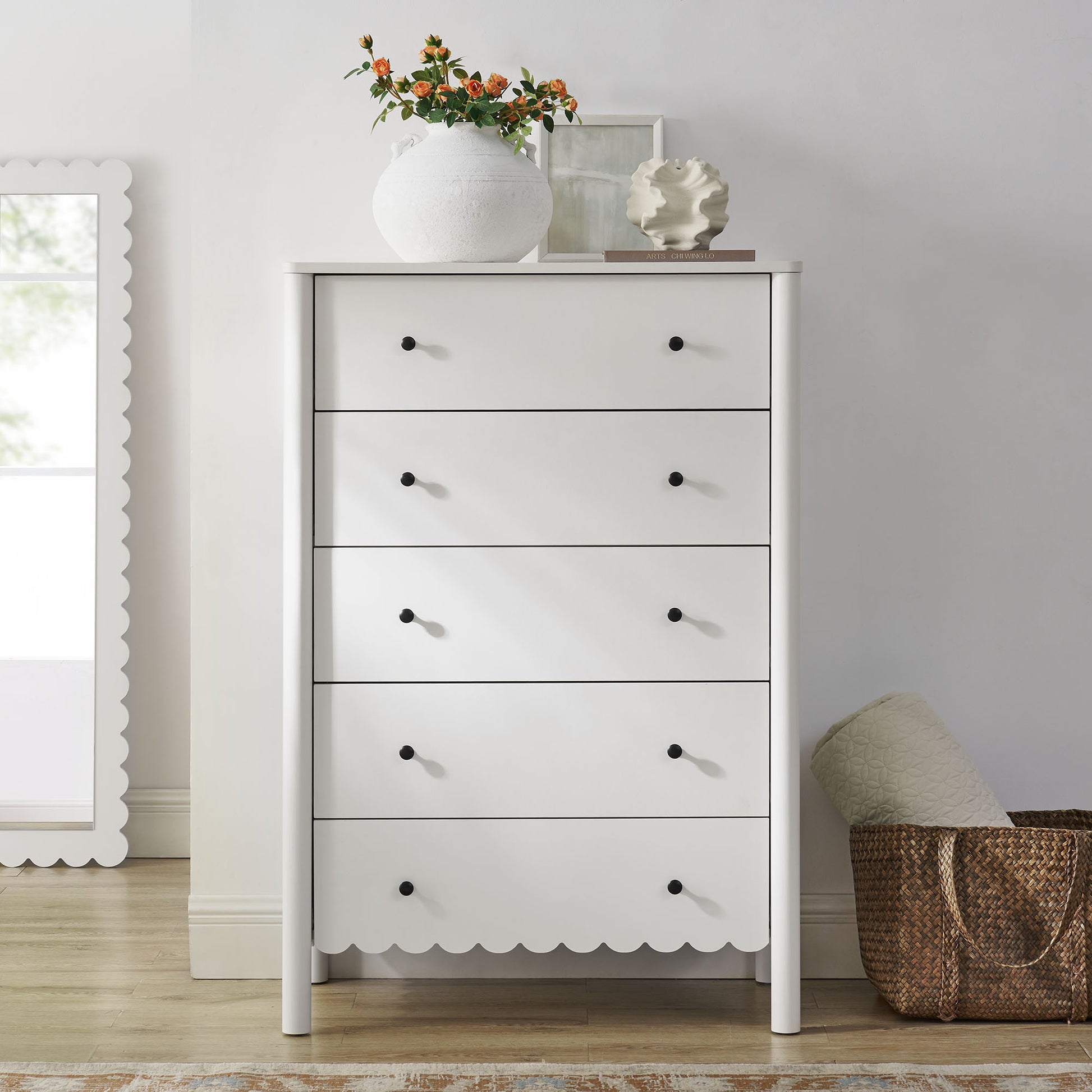 White painted wooden tall chest with five drawers, scalloped apron, black knobs, with a vase of orange flowers on top.