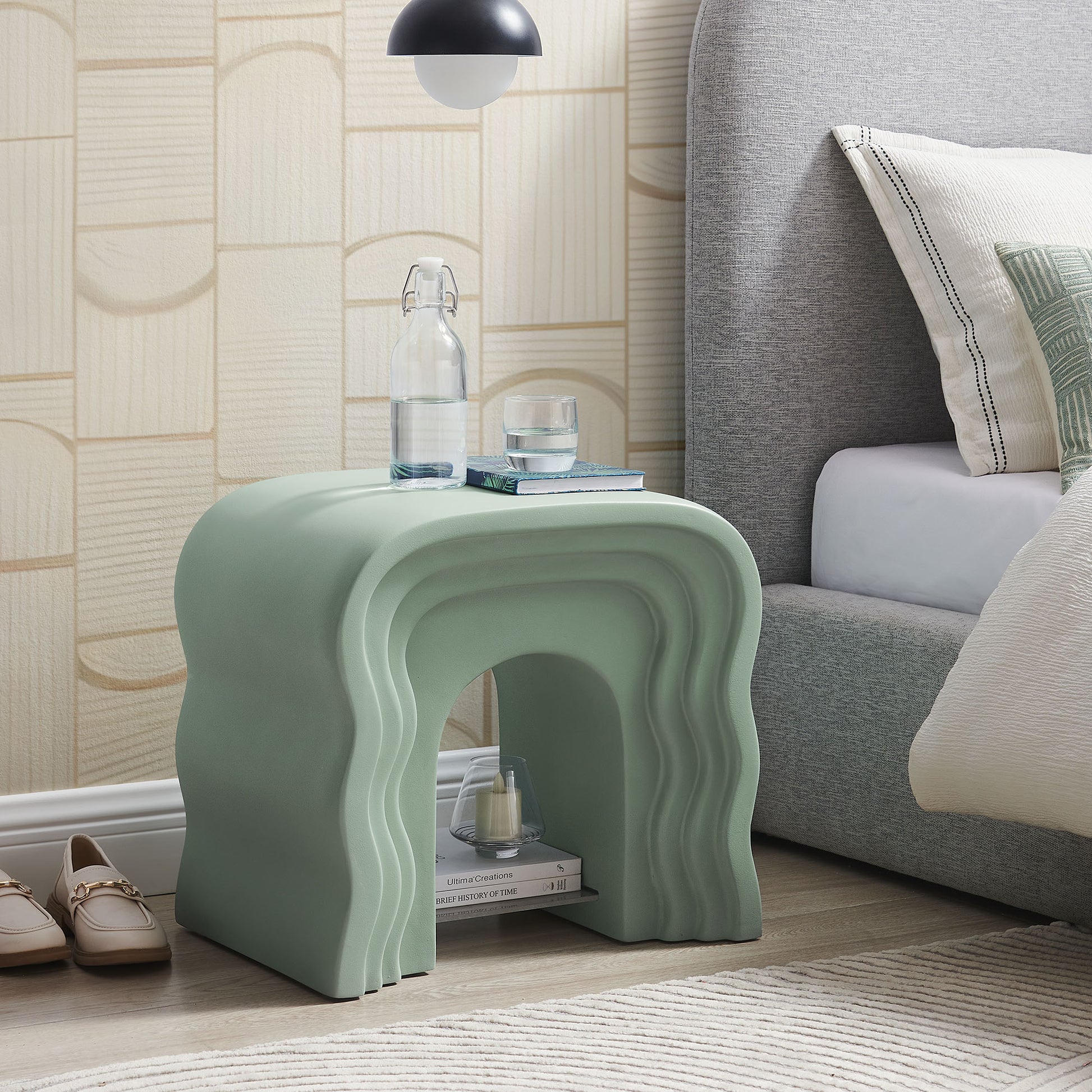Mint-green curved side table with stacked arches and hollow center, placed beside bed with bottle and glass on top.
