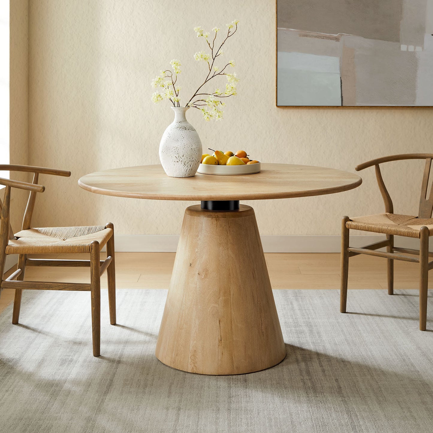 Round natural wood dining table with conical pedestal, two wooden chairs, vase with pale branches, plate of fruit in a neutral room.