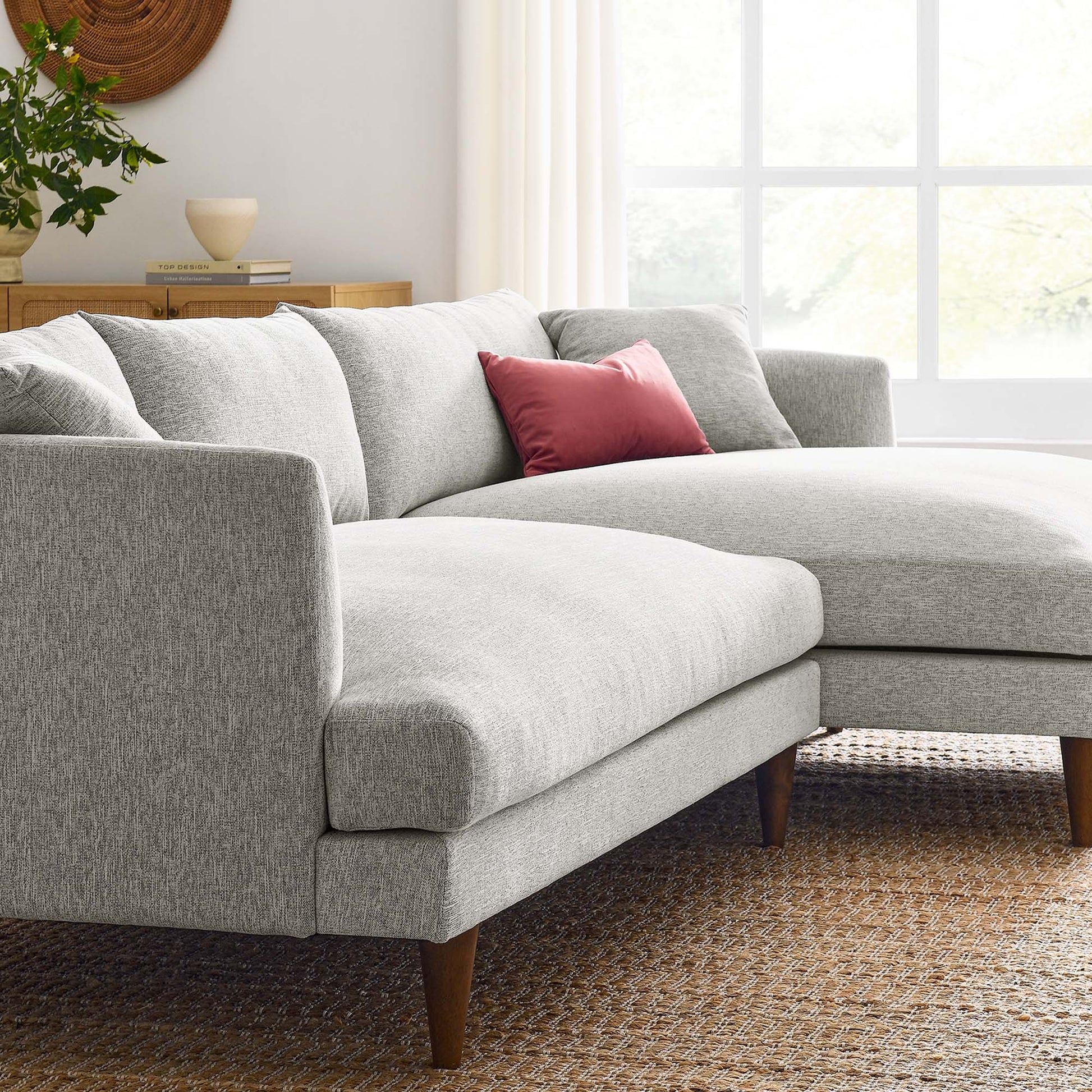 Light gray fabric L-shaped sectional with wooden legs and a pink throw pillow in a bright living room on a woven rug.