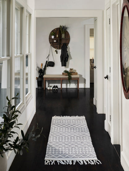 White geometric patterned runner rug with fringe on dark hardwood hallway; bench, circular wood wall shelf, coats and shoes