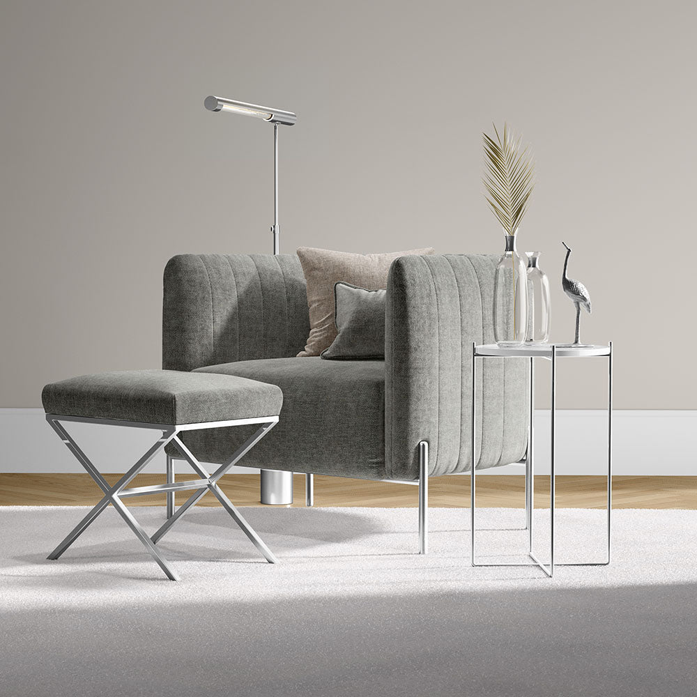 Grey curved lounge chair with matching ottoman and chrome X-frame stool, beside glass-top side tables and modern decor.