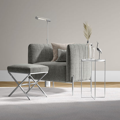 Grey curved lounge chair with matching ottoman and chrome X-frame stool, beside glass-top side tables and modern decor.