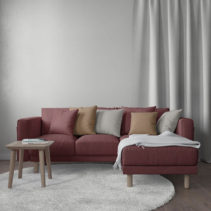 Maroon L-shaped sectional sofa with chaise, multicolor cushions, white throw, light wood legs, round white rug, side table, curtains.