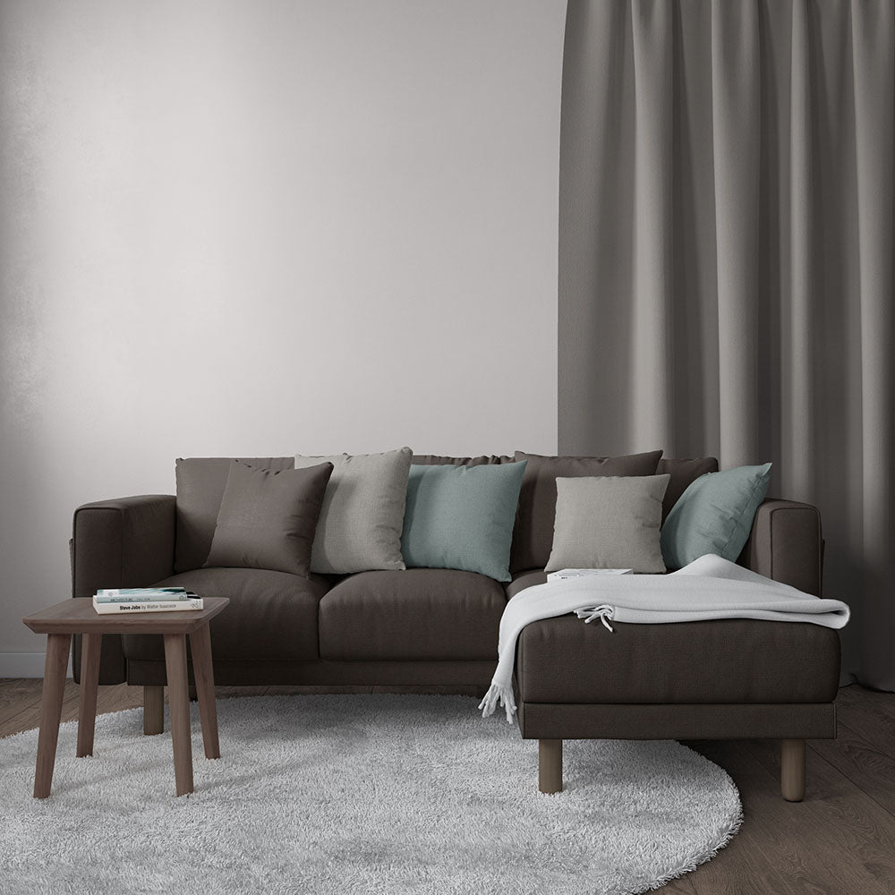 Dark brown L-shaped sofa upholstered in solid brown fabric with chaise, teal & grey cushions, round wood coffee table, white throw blanket, grey curtains.