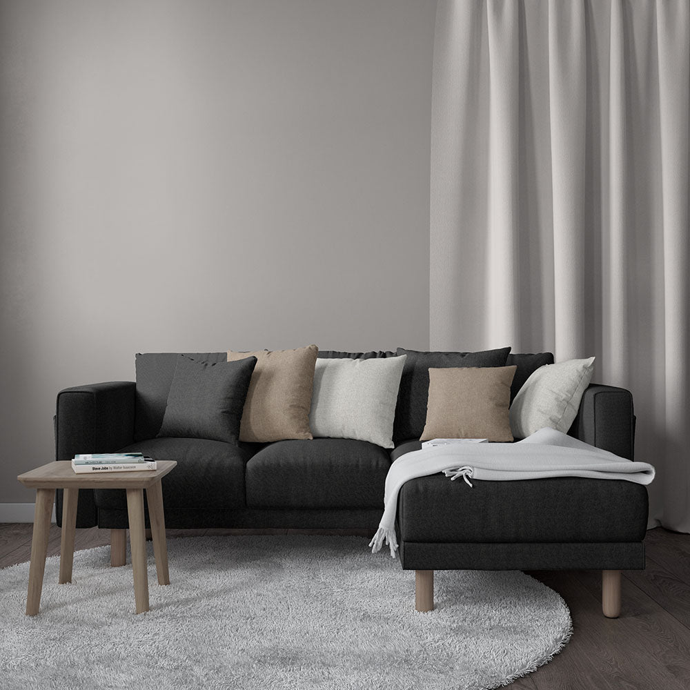 Sectional sofa upholstered in charcoal solid fabric with beige, taupe and white cushions, light gray throw, wooden coffee table and soft curtains.