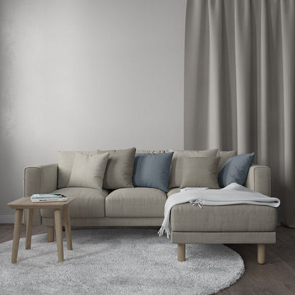 Beige fabric sectional with chaise, blue and beige cushions, light wood legs, grey rug, small wooden table, grey curtains.