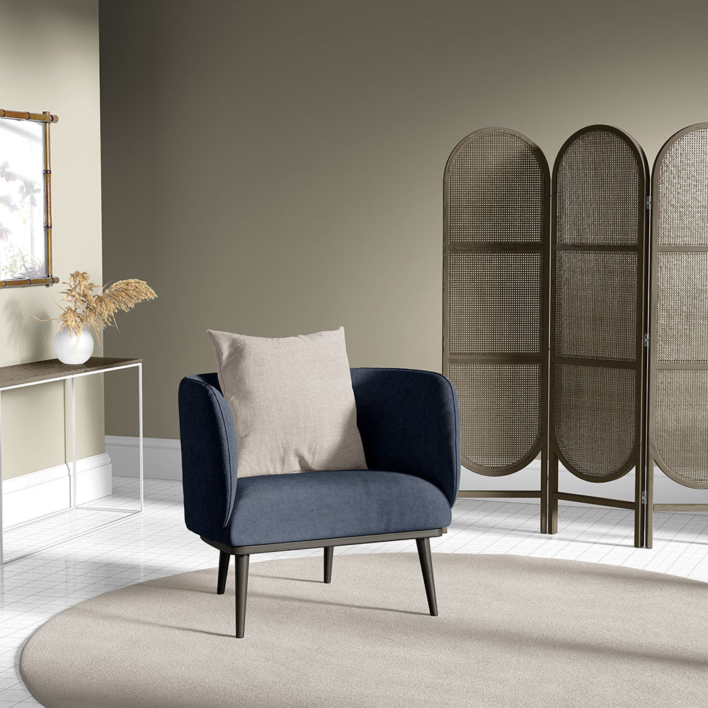 Dark blue velvet armchair with beige cushion, mid-century legs, in a neutral living room with a woven screen divider.