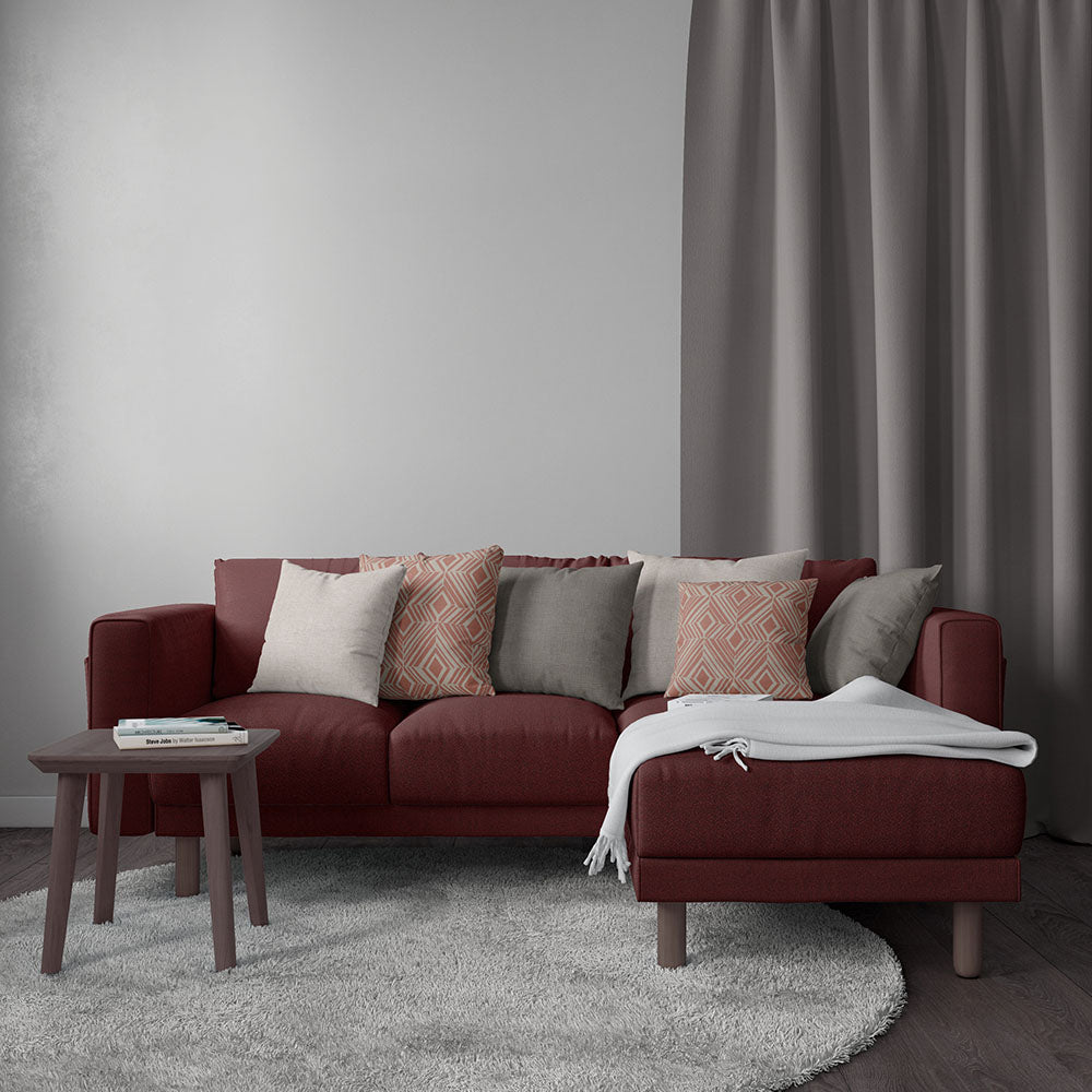 Maroon fabric sectional with chaise, mixed cushions, grey throw, round light-grey rug, small wood side table, grey curtains.