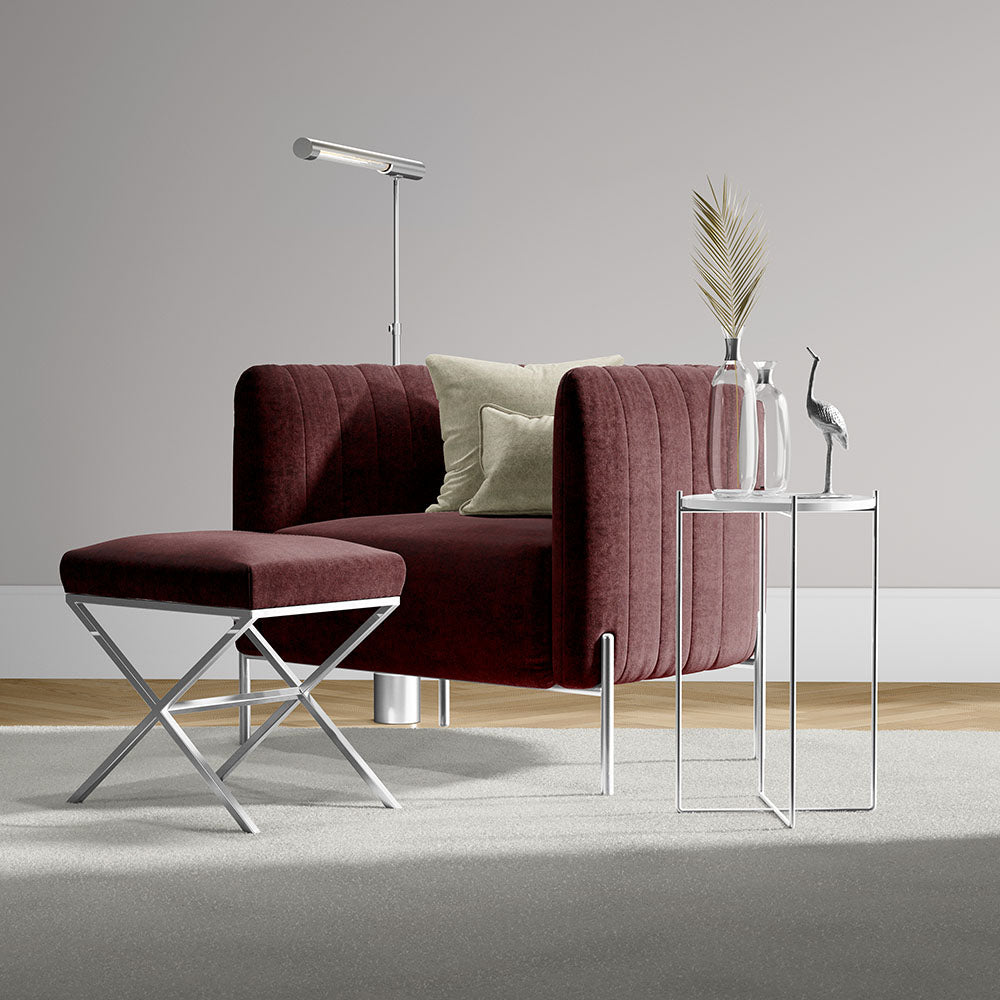 Maroon velvet armchair with ottoman and chrome legs, beside glass side table with vase, dried palm and a metal bird statue.