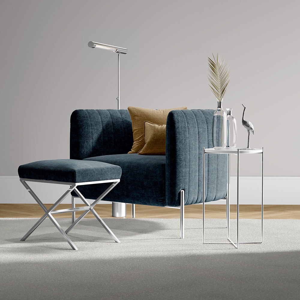 Dark blue velvet accent chair with ottoman on chrome X-frame legs, glass-top side tables, a mustard cushion, floor lamp.