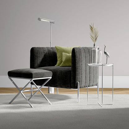 Dark gray velvet armchair with ottoman, chrome X-frame legs, glass side tables, floor lamp, and green cushions.